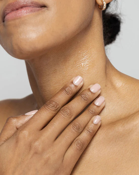 Close-up of a person's neck and hand with light pink nail polish on a neutral background appling Kine-C to the neck area to smooth the appearance of fine line and wrinkles